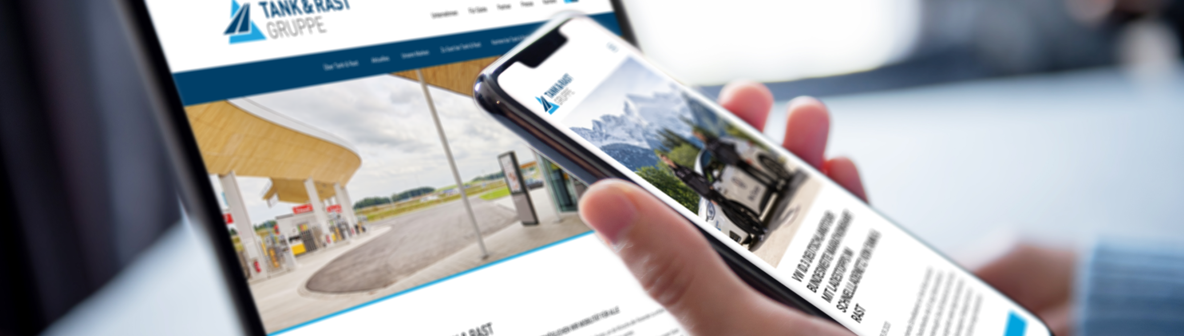 Eine Person hält ein Smartphone in der Hand auf dem ein Pressebeitrag der Tank und Rast zu sehen ist. Hinter dem Smartphone steht ein Laptop auf dem Tisch auf dem die Startseite der Tank und Rast Webseite geöffnet ist.