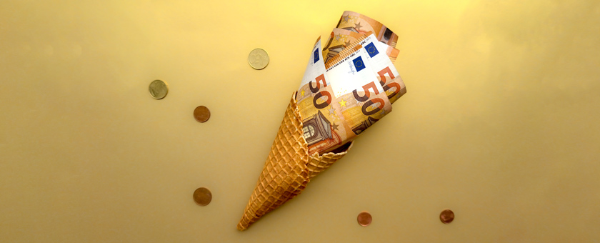 Fünfzig Euro Scheine sind in eine Eiswaffel gewickelt und schauen oben raus. Um die Eistüte mit den Geldscheinen liegen Münzen verstreut.