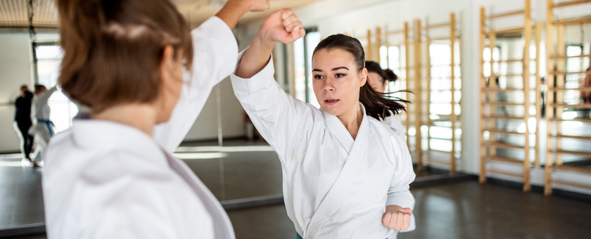 Zwei Frauen stehen sich in weißer Kampfkunstkleidung in einem Trainingsraum gegenüber. Die Frau im Zentrum der Fotografie wehrt einen Angriff ihrer Gegnerin ab.