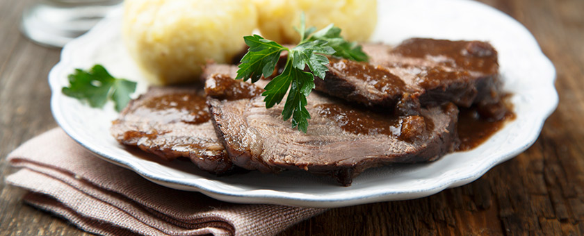 Auf einem Tisch steht ein Teller mit drei Scheiben Sauerbraten und Kartoffelklößen.